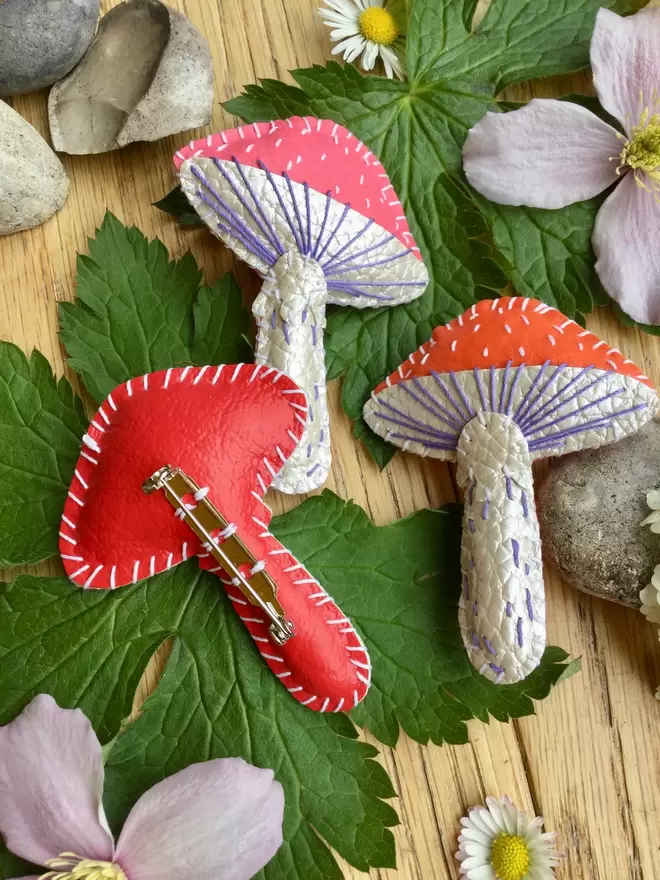 Three hand stitched faux leather toadstool brooches, in red, pink and orange laying amongst leaves and flowers on a wooden surface. The red toadstool brooch has been upturned to display the brooch back on the red reverse side.