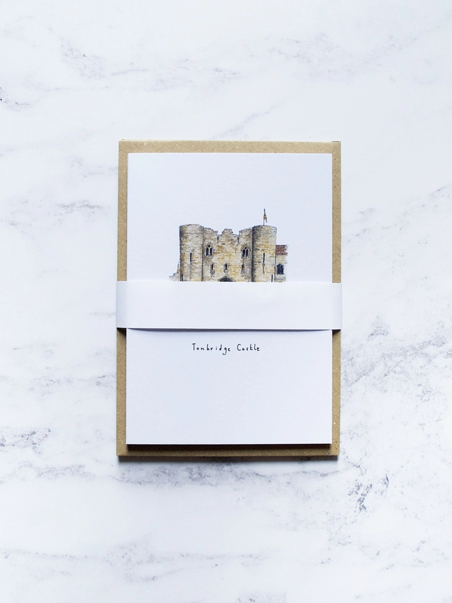Watercolour illustrated pack of Tonbridge cards on A6 white card, with brown kraft paper envelope behind. The top card is Tonbridge Castle, Tonbridge, but is slightly obscured from view by the white band holding the pack together. The background of the image is a pale white marble. 