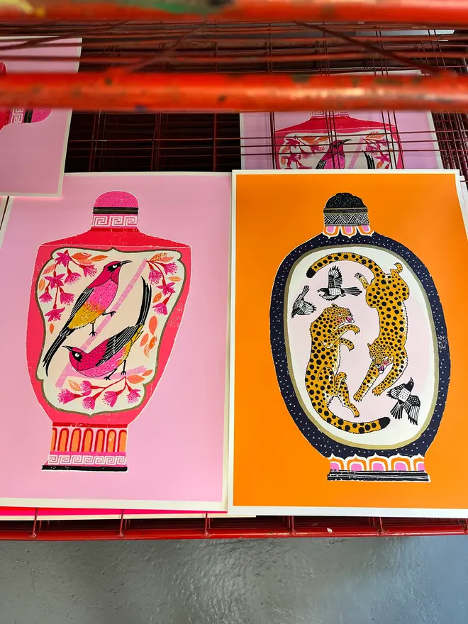 Sunbirds Snuff Bottle Screenprint Sunny Sunbirds, a colourful pink print of a pair of birds in a vase on a pink background. It is accompanied by another orange print on a studio rack. 