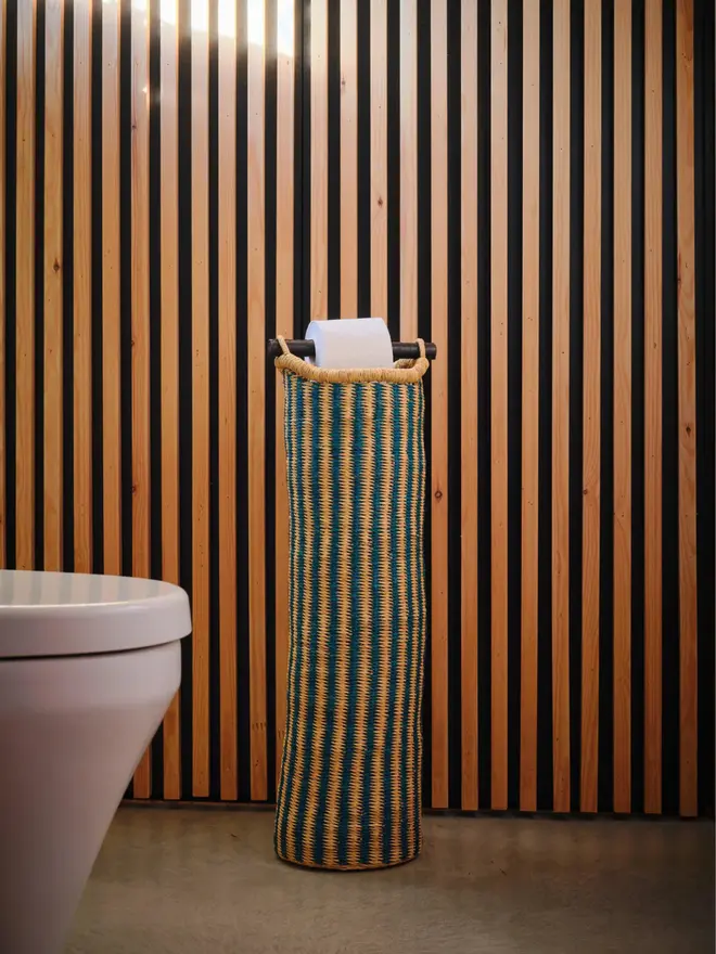 Ghanaian Hand Woven Tubular Toilet Roll Holder, a toilet roll holder sitting in a bathroom. It sits in front of a wooden backdrop. 