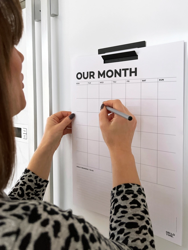 Lady writing on a Year Wall Planner hung up on a white fridge.