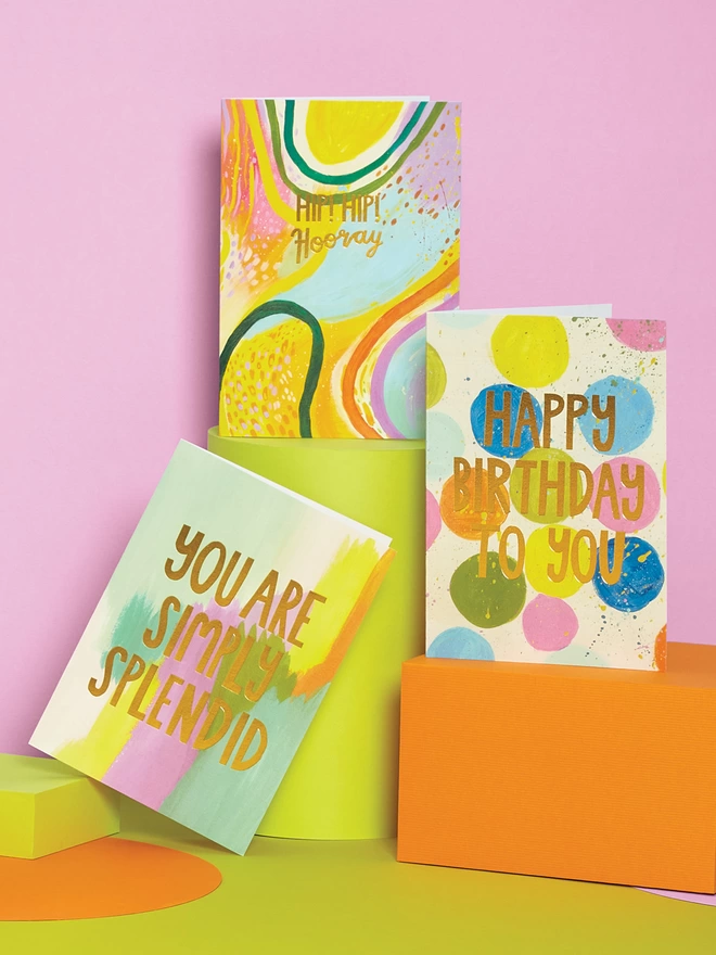 Three painterly cards from the Raspberry Blossom ‘Canvas Creations’ greeting card collection sit on orange plinths in front of a pink and green background