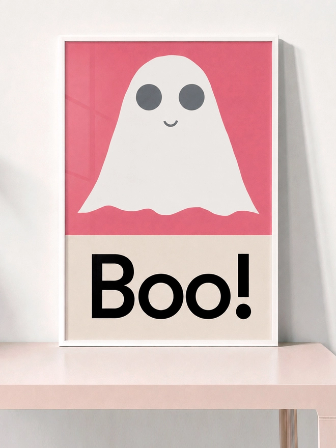 A framed print with a white ghost and gray eyes on a pink background. Below the ghost, the word "Boo!" is printed in bold black letters on a cream background. Perfect for Halloween.