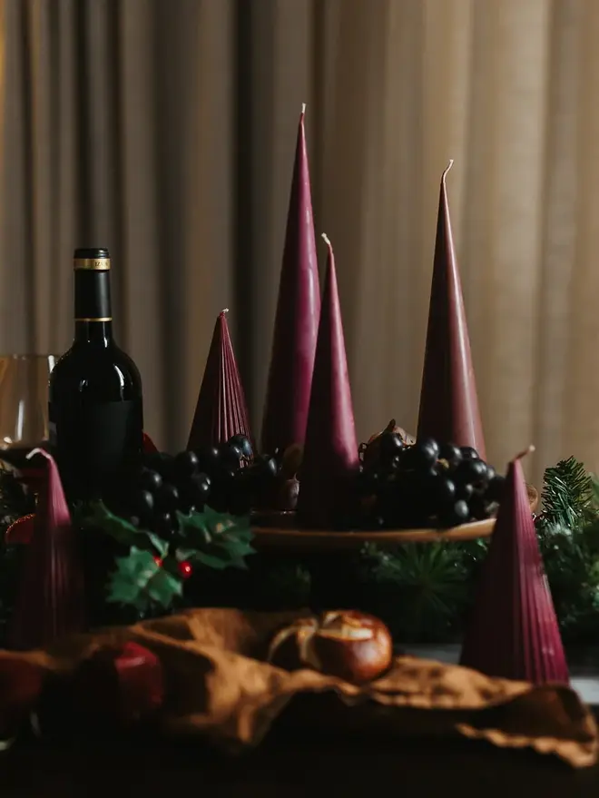 Burgundy Christmas Tree Cone Candles (Set Of 3). A long festive table lined with burgundy cone candles, a bottle of red wine, grapes and pine garland, with fairy lights glowing behind. Tall and short candles together give the tablescape height and drama for holiday entertaining.