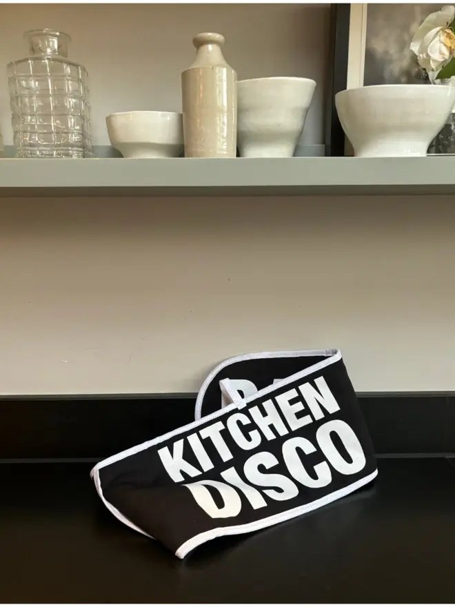 Black oven glove with white text that says KITCHEN DISCO sat on black counter with beige shelf above that has ceramics and glass objects and a framed black/white photo sat on it