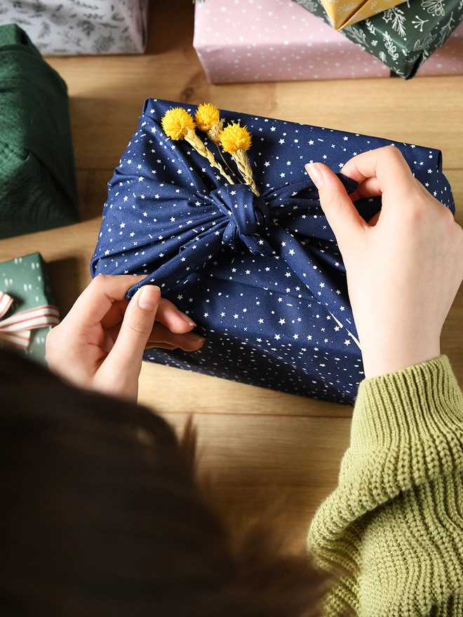 A gift is being wrapped in navy blue cotton fabric wrap with a white star design, and a small posy of yellow flowers is being tucked into the knot.