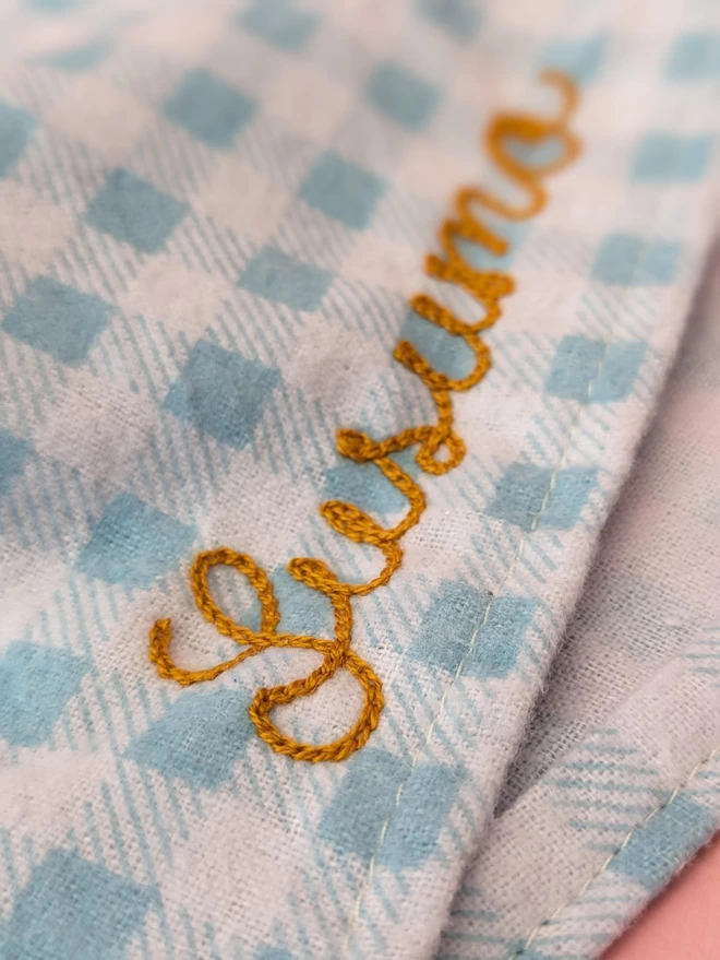 Powder blue and white checkered dog bandana close-up  shot, with gold embroidered personalisation reading 'Susumo' on a pink background