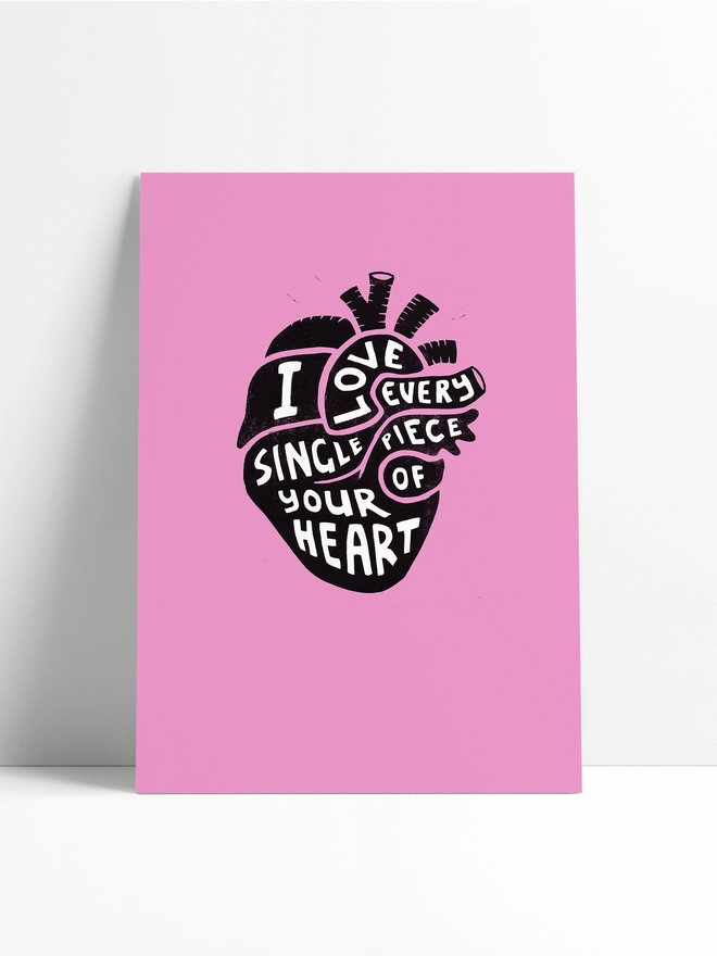 A pink Woodism print, unframed, in a limbo background, featuring a black anatomical heart with white typography which reads: I Love Every Single Piece Of Your Heart.