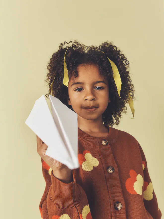 Close crop of a child with yellow hair ribbons holding a paper plane towards the camera, wearing a Studio Koter chestnut floral intarsia cardigan.