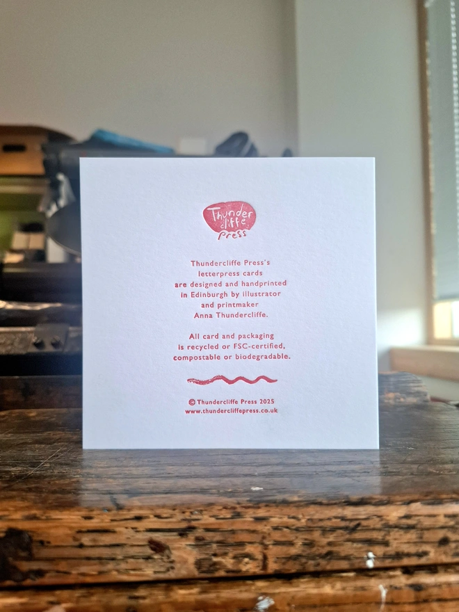 A handprinted greetings card standing open on a wooden surface, showing the text on the back, printed in pink ink.