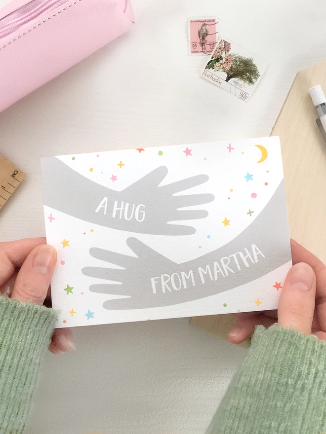 A friendship greetings card with a hugging arms design and personalised wording that reads "A hug from..." is being held above a white desk.