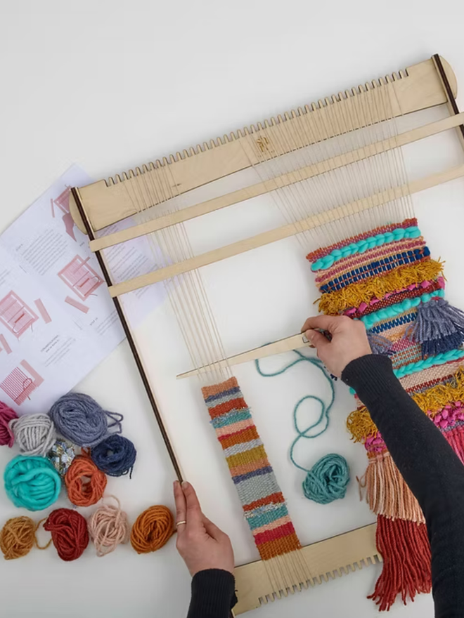 large weaving kit. a large loom on a white surface, with yarn in various colours and a person in the middle stages of the weaving process.