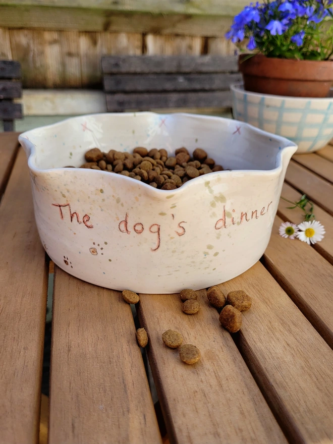 ivory ceramic dog bowl with 'the dog's dinner' written on the side with brown green and blue splatters and pawprints on a table with dog biscuits in