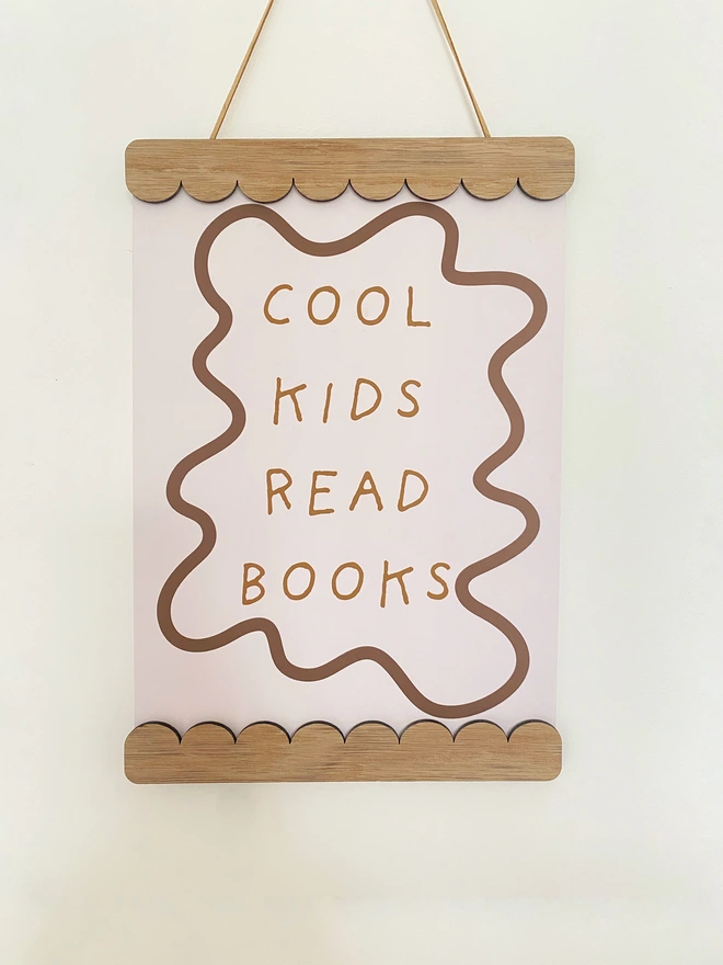A wooden magnetic picture frame, strung with vegan suede. Hung on a wavy wooden wall dot. Displaying a colourful stripy kids print that says cool kids read books 