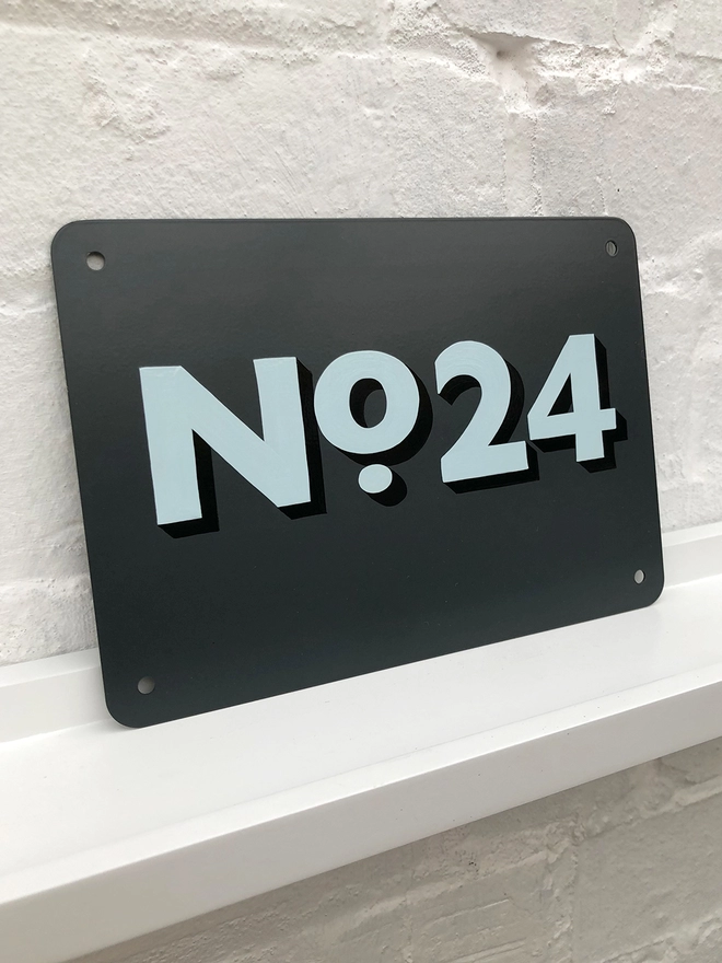 Hand painted pale blue and black house No. 24 on an anthracite grey metal plaque against a white brick wall. 