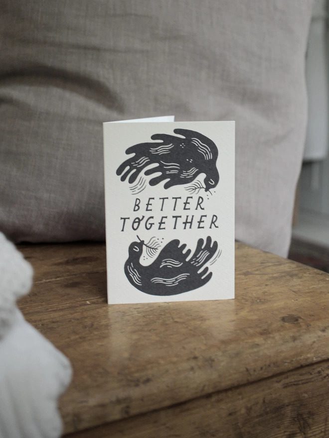 Black and white greeting card with illustration and the words better together written on it stood up on wooden surface with a grey cushion behind