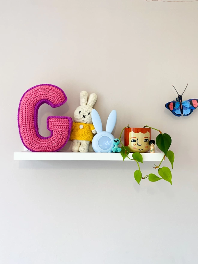 Crochet Cushion shaped like the letter G in Pink and Magenta, on a child's shelf