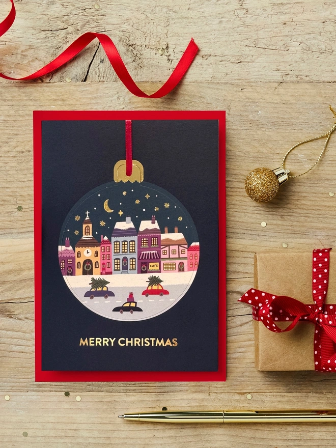 Christmas pop-out bauble card with snowy town illustration on it sat on a wooden desk with red and gold Christmas decorations.