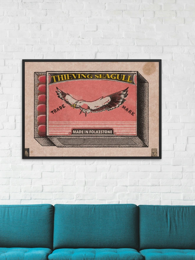 
A close-up photograph of a limited edition A2 screenprint. The print depicts a retro-style matchbox illustration in pink, black, and gold. A large, realistic seagull with a mischievous expression and an open beak is flying across the center. The words "THIEVING SEAGULL" are at the top, and "TRADE MARK" is on either side of the seagull. A banner at the bottom says "MADE IN FOLKESTONE." The background is a solid pink with a slight gold texture. The matches on the side are green and heart-shaped. Below the main image, the print is signed and numbered 3/13. The entire image has a grainy texture, like a vintage screen print.