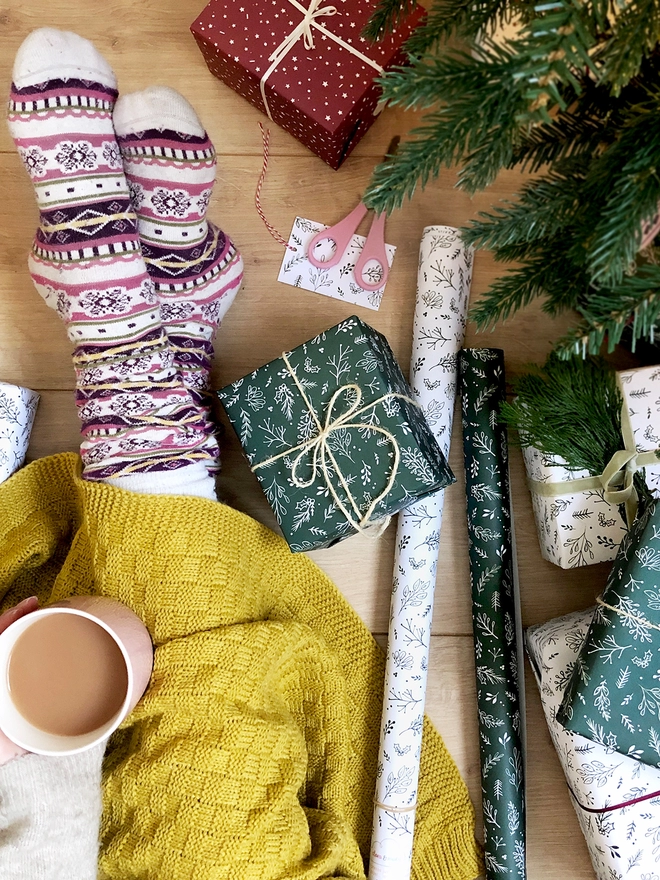 Several gifts wrapped in green and white greenery wrapping paper are laid on a wooden floor beside someone sitting drinking tea.