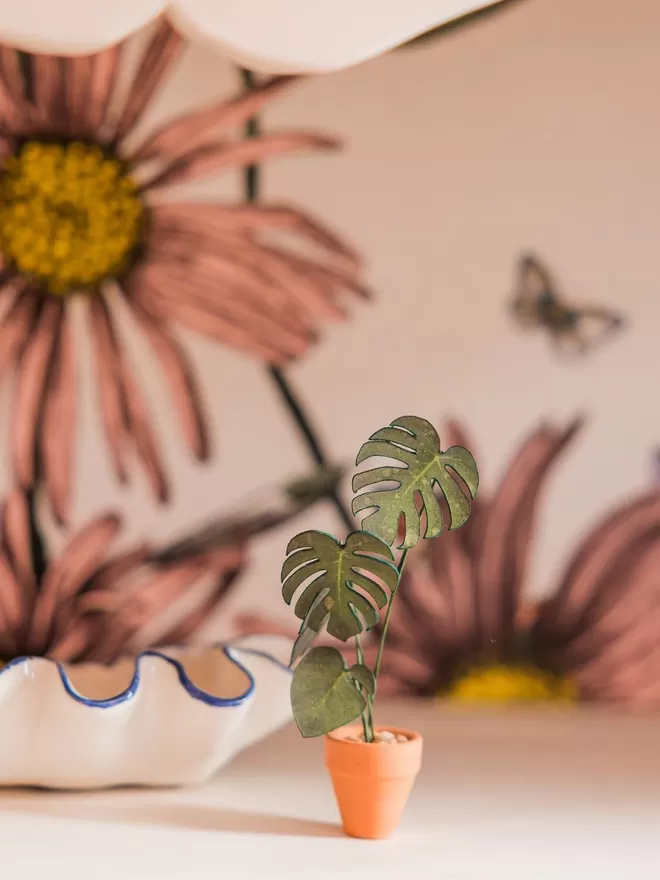A miniature replica Monstera Deliciosa paper plant sat on a wooden tray with succulent plants in the background