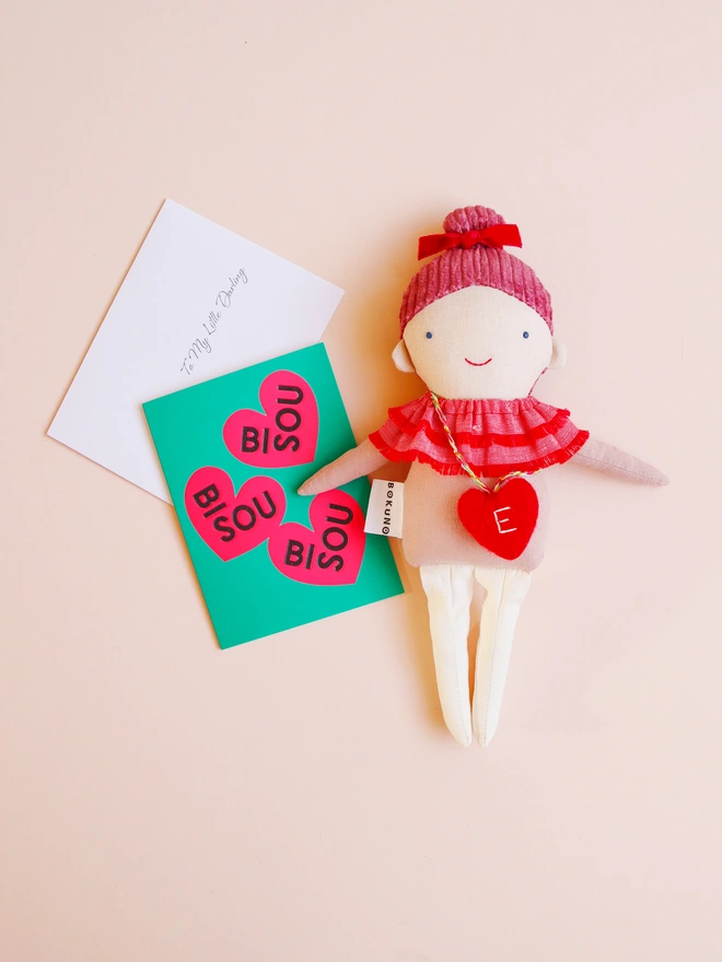 Handmade cloth doll with a pink corduroy hair with a red velvet bow and a layered red ruffle collar and pink outfit, wearing a small red felt heart bag with the letter “E”