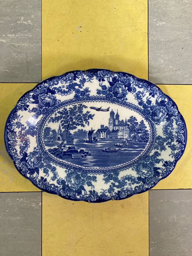 Classic blue and white patterned plate featuring a castle and river with boats sailing along. A Jet2 plane flies high in the sky.