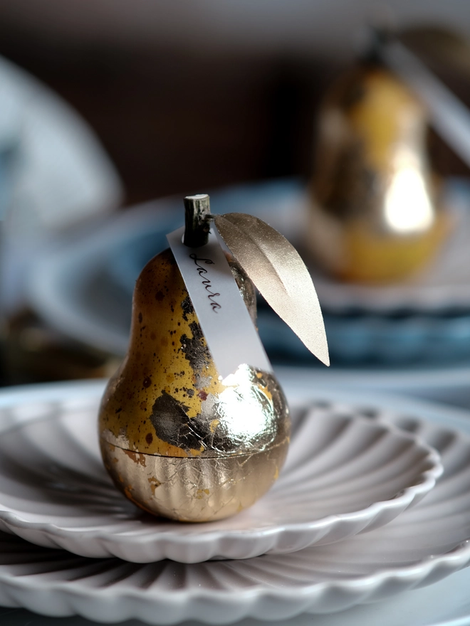 Gilded vintage-style Christmas pear ornament opened to reveal a small surprise inside, shown on a festive Christmas table setting with white plates and ribbon.