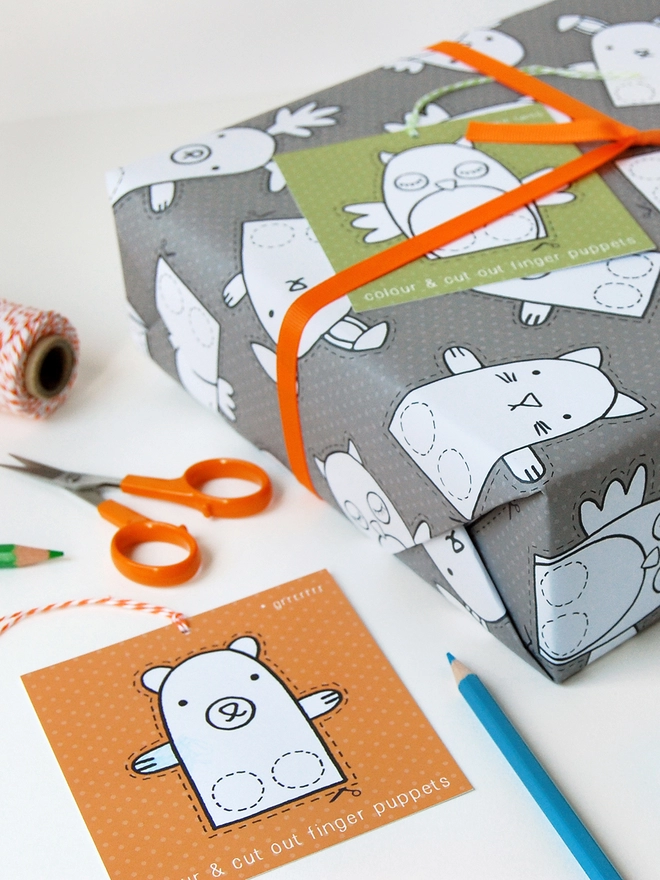 A gift wrapped in grey wrapping paper that is covered in drawings of animal finger puppets that can be cut out and coloured lays on a white desk.