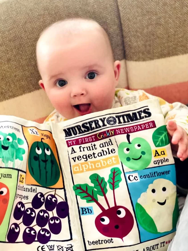 Baby holding the Fruit and Vegetable Alphabet Crinkly cloth newspaper smiling