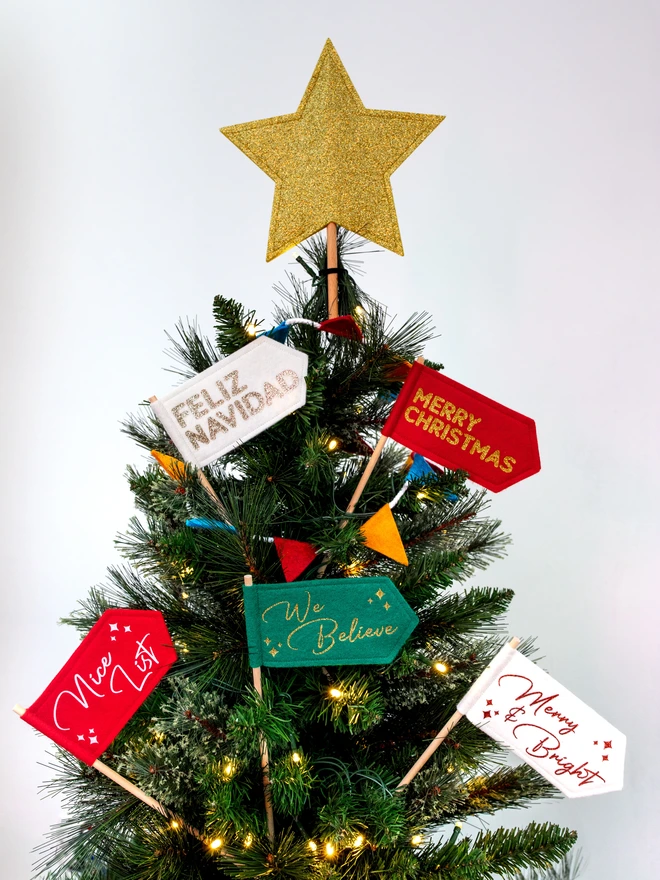 A range of christmas flags in a variety of colours and messages displayed on a christmas tree with a glitter gold star on top The flags are made from felt and the text is written in a mix of bold and cursive glitter letters