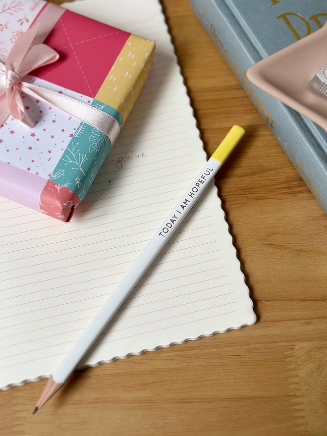 A white pencil with a yellow end and the words "Today I Am Hopeful" along the side lays on an open lined notebook, which rests on a wooden desk.