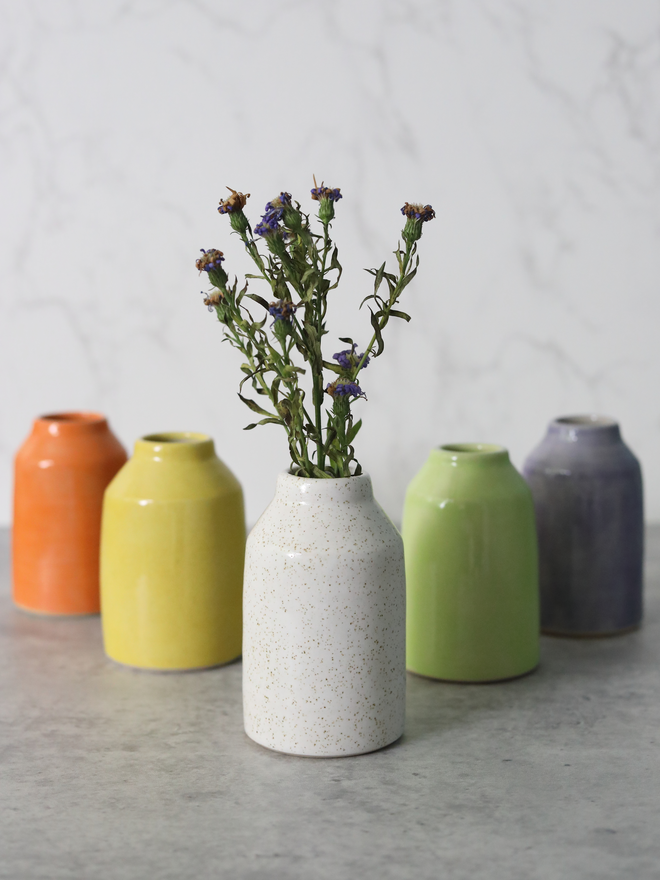 A speckled white handmade ceramic bud vase holding dried purple wildflowers, photographed in focus in the foreground with four colourful vases — orange, yellow, spring green, and slate — softly blurred behind it on a grey marble surface