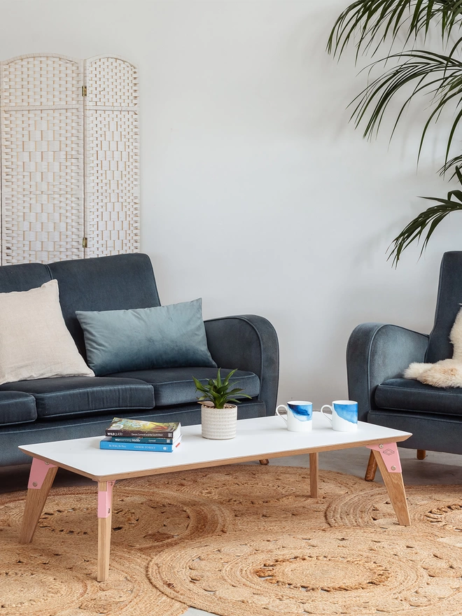 A stylish and minimalist low coffee table with white Fenix top, pink coloured steel brackets and solid oak legs on a jute rug surrounded by blue chairs