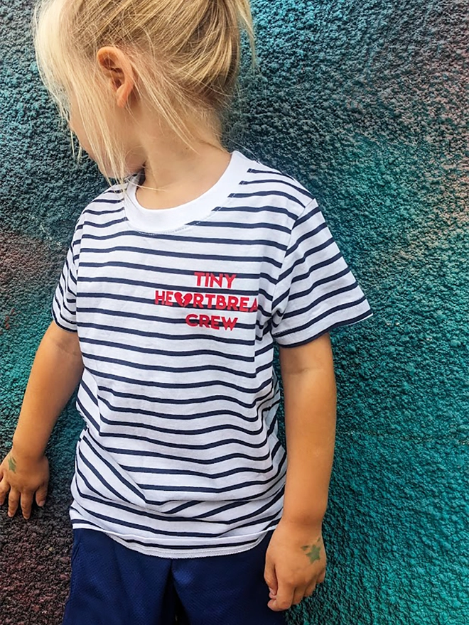 Child wearing a navy and white stripe t-shirt with red Tiny Heartbreak Crew slogan.