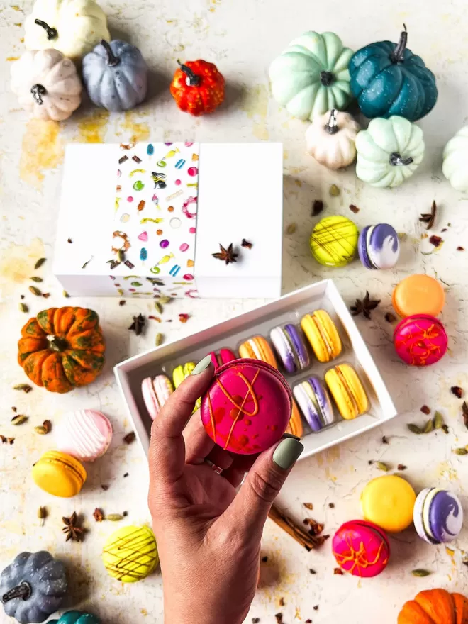 a person's hand holding a hot pink & orange macaron in front of a box of colourful autumn macarons surrounded by colourful pumpkins on a table 
