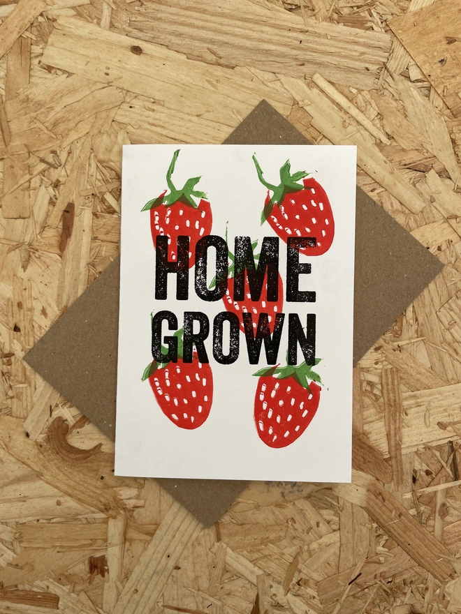 A white greeting card on a wooden surface shows  illustrated red strawberries with green leaves surrounding bold black text that reads “HOME GROWN.”