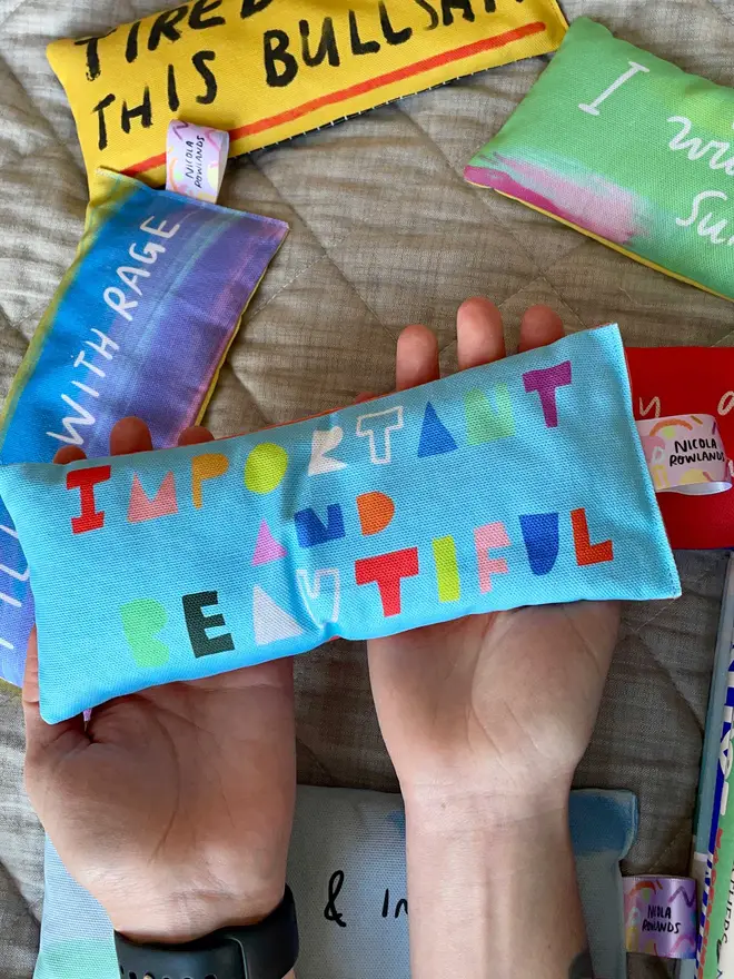 Holding in hands outstretched a light blue lavender eye pillow which reads in brightly coloured letters IMPORTANT AND BEAUTIFUL