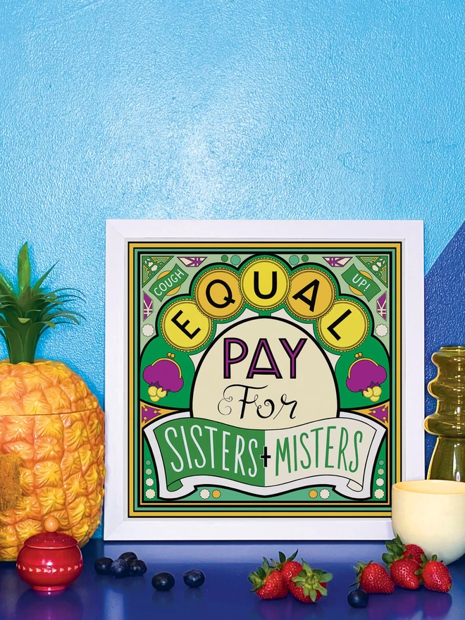 Equal Pay for Sisters and Misters is written over this green and yellow illustration. In the corners “Cough” and  “Up!” appears in banners at the top corners. It is in a white square frame propped against a wall painted light and dark blue, on a shiny blue cabinet. Next to the frame is a plastic pineapple ice bucket, a small wooden red pot a handful of blueberries, a yellow glass vase, a small yellow ceramic pot and 4 strawberries. 