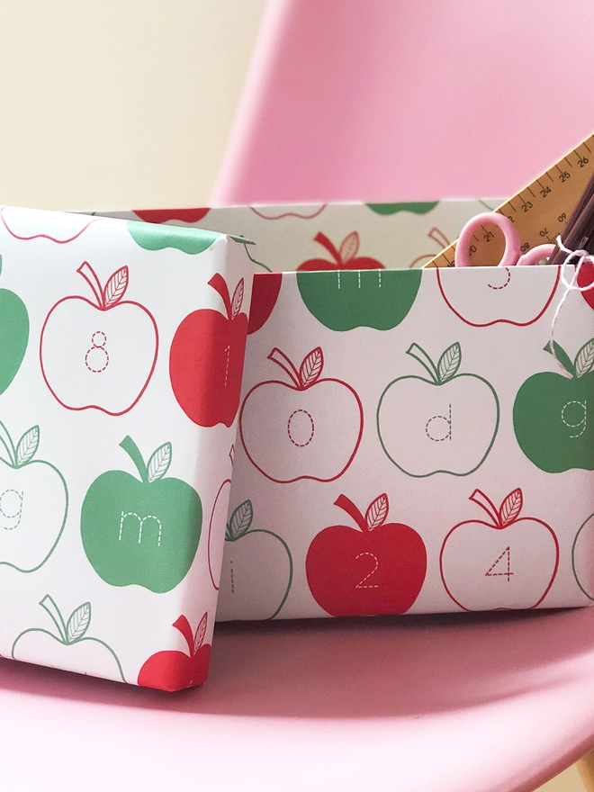 A gift wrapped in red and green apple wrapping paper, with letters and numbers inside each apple, rests on a pink chair.
