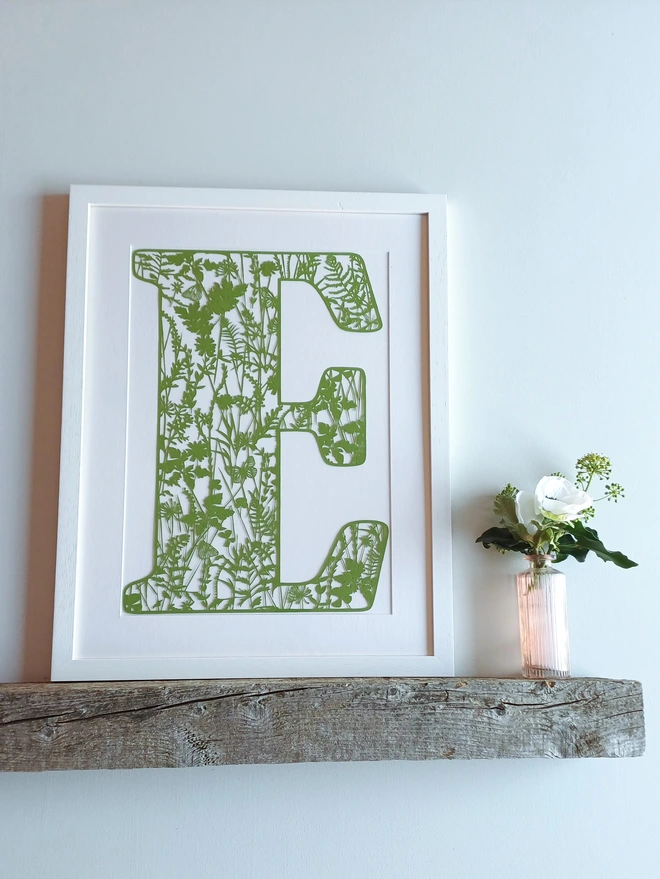 Summer Papercut Letter E in olive green, framed in narrow white frame on wooden shelf