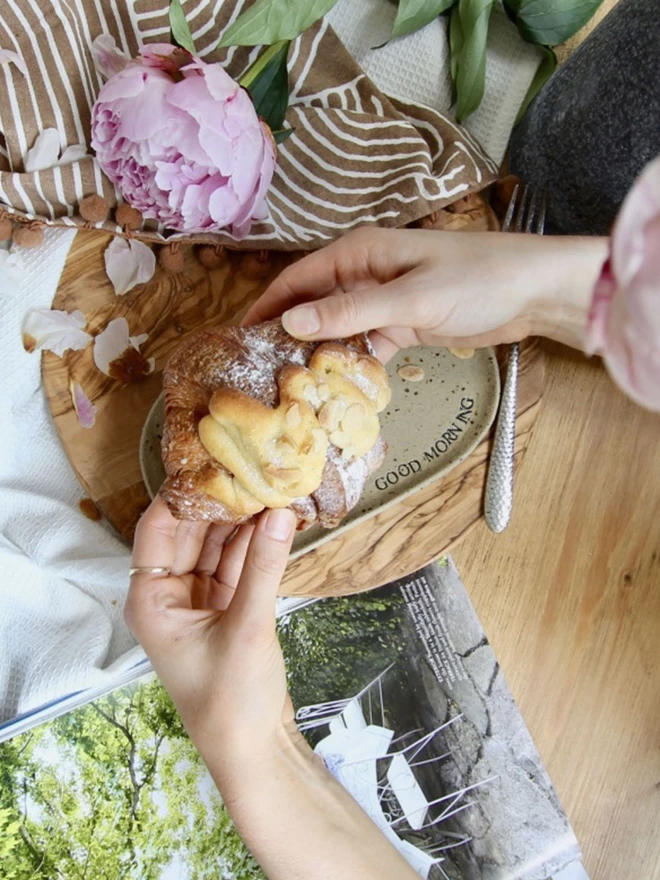 hands holding an almond croissant above an oval ceramic plate embossed with the words 'Good Morning'