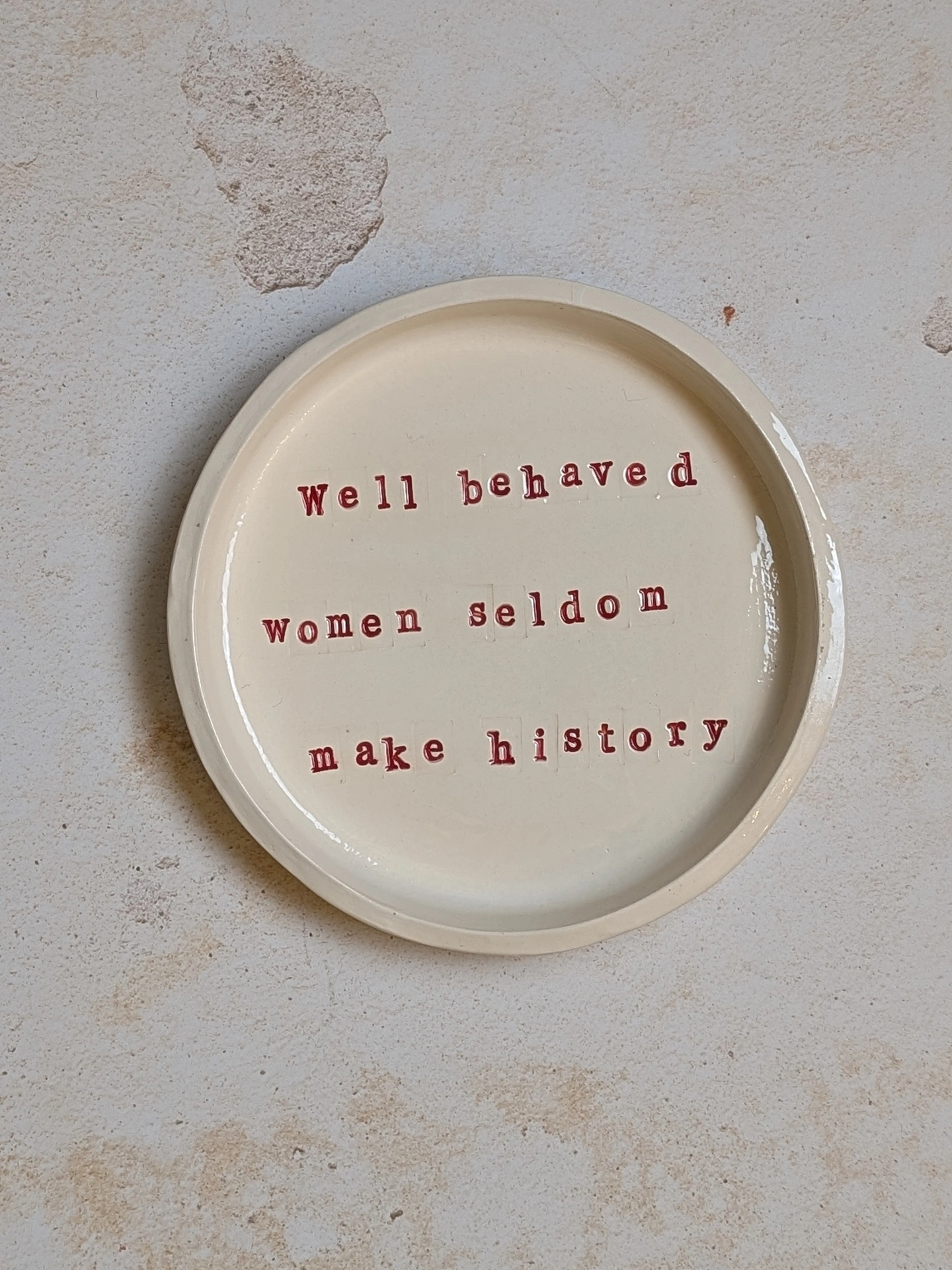 Well behaved women seldom make history a ceramic dish sitting on a grey surface. It is inscribed with a message in red writing. 