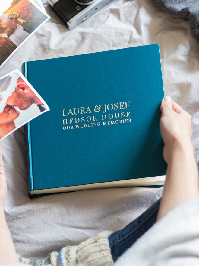 a woman is looking through her wedding guest book whilst sitting on her bed. The guest book is blue and has names and a wedding venue printed on the front of it in gold lettering.