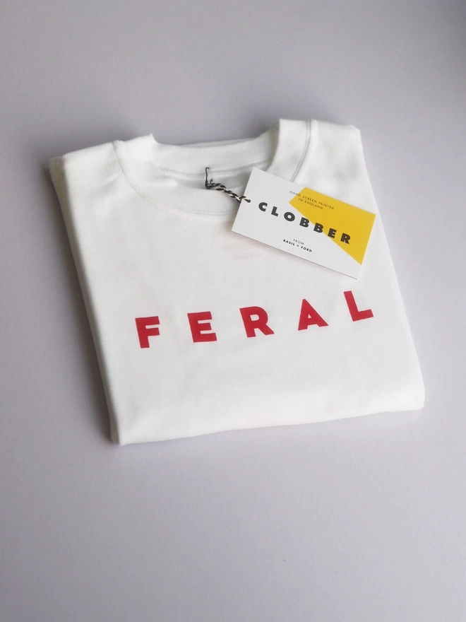 photograph of a white kids t-shirt folded with the design FERAL over the top in red writing. Very cool trendy t-shirt with Basil & Ford Clobber written on the  retail tag