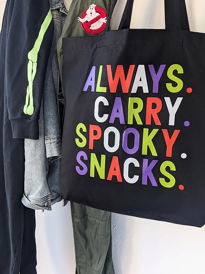 A black tote bag with multi coloured Always Carry Spooky Snacks slogan.