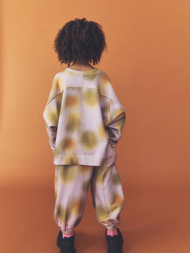 Back view of a child standing against a burnt orange backdrop, showing the yoke seam and side split of the Studio Koter Riverson top in Hazy Lilac, worn with matching cuffed print joggers.