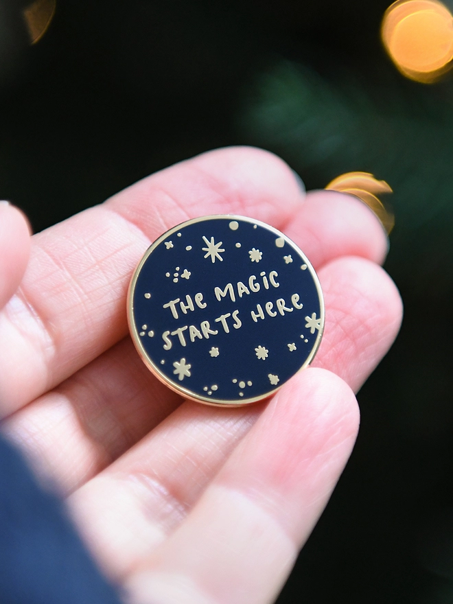 A navy blue and gold enamel pin badges with a starry design and the words "The magic starts here" rests on an open hand.