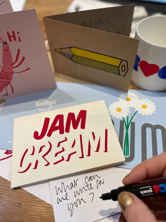 A hand is poised over a pile of jolly greetings cards, ready to write a message in energetic handwriting. Jam Cream is the main card, screenprinted in a cool font, red ink on cream card. Other cards are around, a yellow pencil a pink prawn, flowers in a vase.