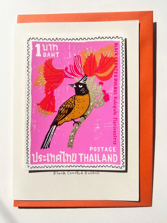 Bird Stamp Card Thailand Black Crested Bulbul, a white card with a colourful bird design in the centre on top of an orange envelope on a white surface. 